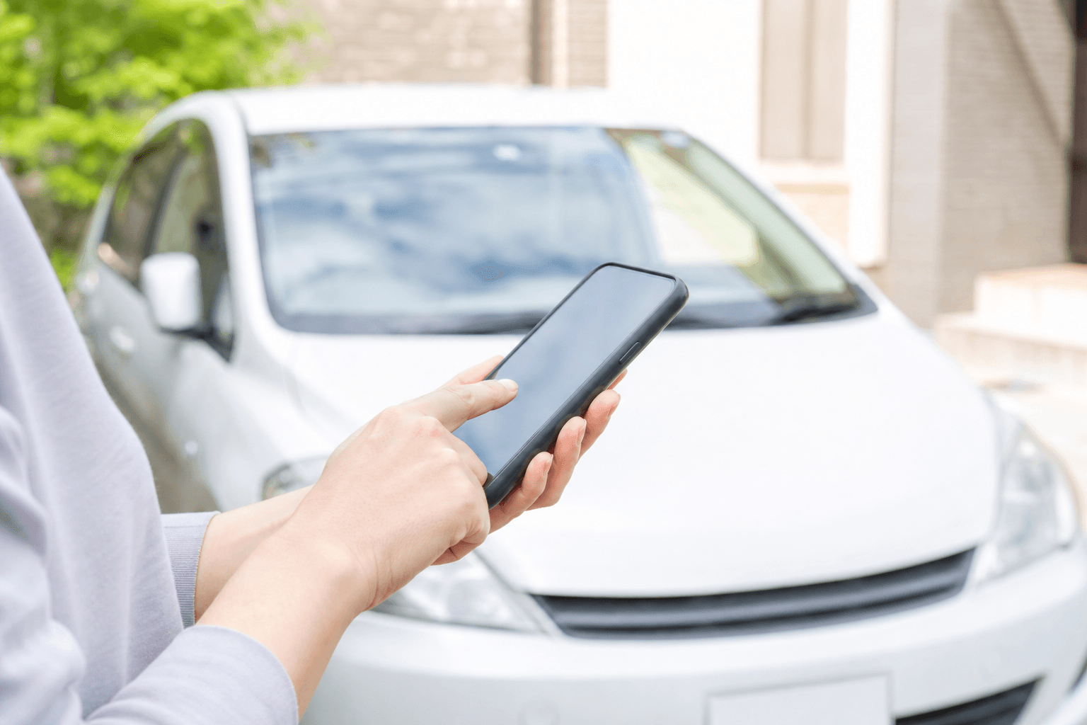 自宅の駐車場で、白いコンパクトカーの前でスマートフォンを操作する手元
