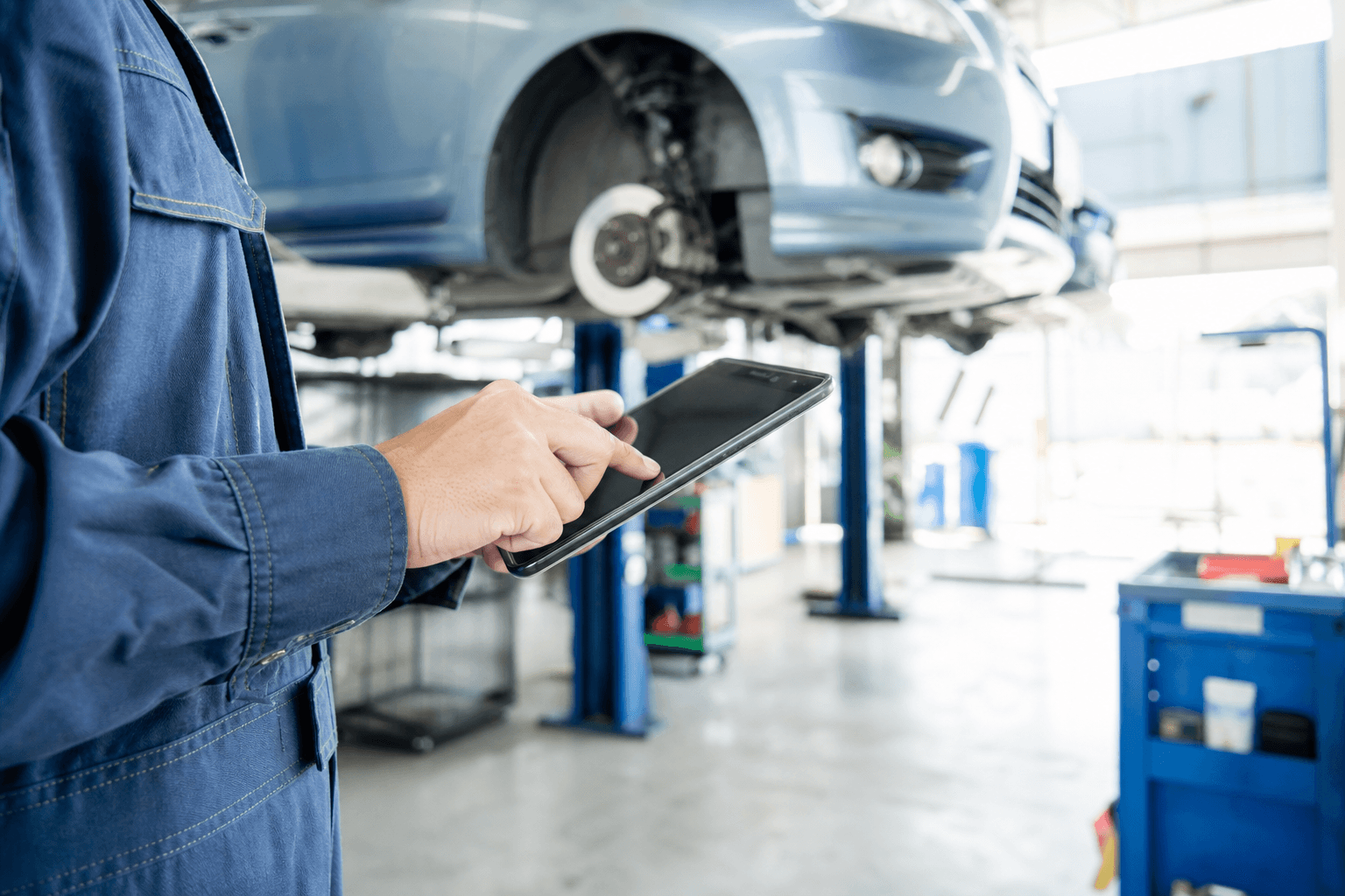 整備工場でタブレットを操作する整備士の手元。奥にはリフトアップされた車