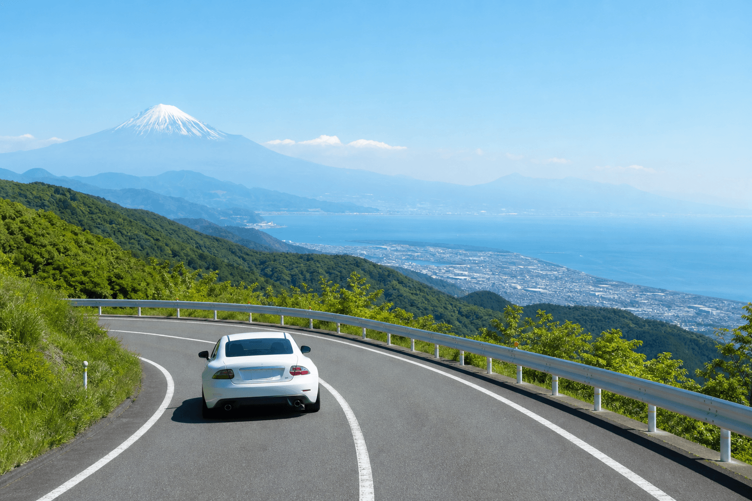 A white sports coupe driving a winding mountain toll road with Mt. Fuji and Sagami Bay in the distance