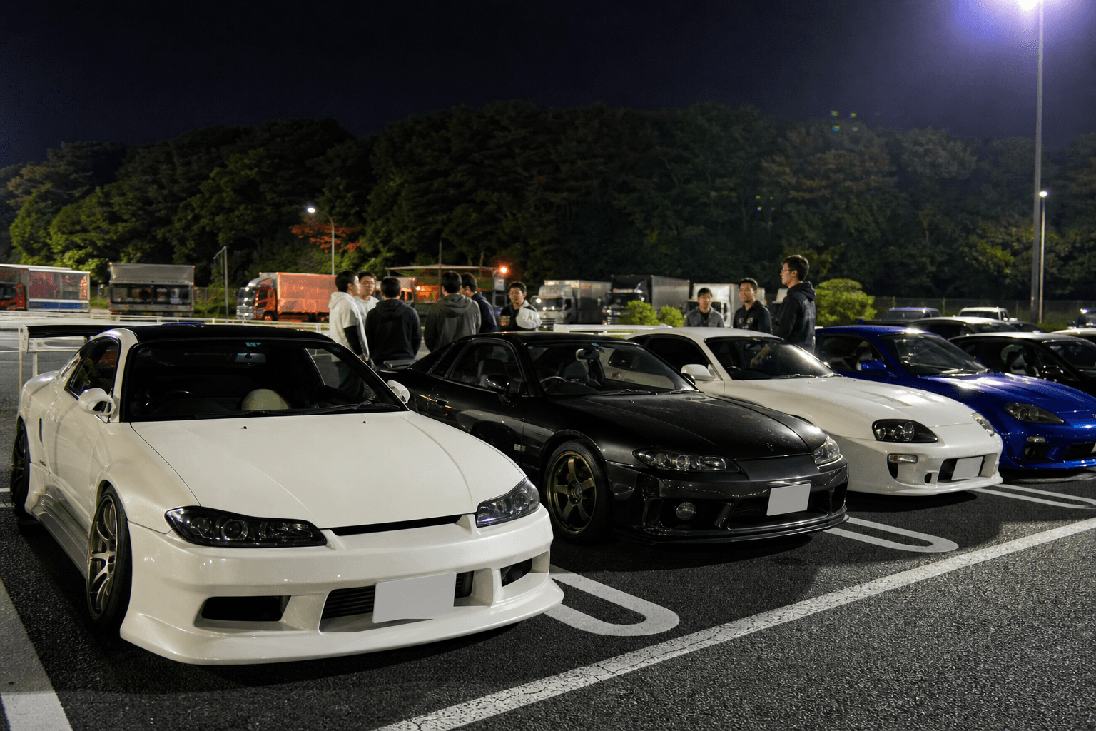 Modified Japanese sports cars lined up at a highway parking area at night while enthusiasts chat